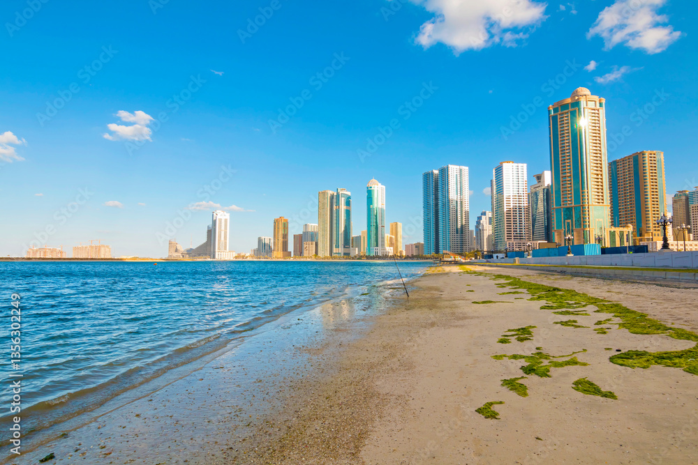 Obraz premium Sharjah city skyline at sunrise with golden light reflecting on modern skyscrapers. Serene beach with gentle waves. Peaceful morning atmosphere in the UAE’s cultural capital.