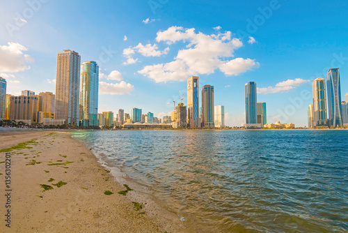 Obraz na płótnie Sharjah city skyline at sunrise with golden light reflecting on modern skyscrapers. Serene beach with gentle waves. Peaceful morning atmosphere in the UAE’s cultural capital. w salonie