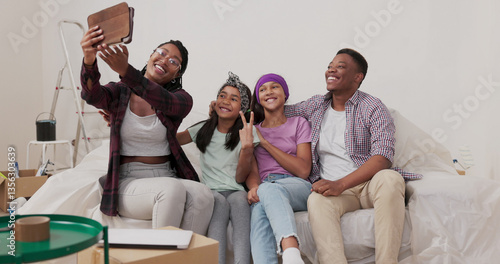 A joyful couple with their two daughters sits on the sofa in the renovated apartment. The woman holds a tablet, trying to capture a selfie with her family, laughing at the unsuccessful shot.