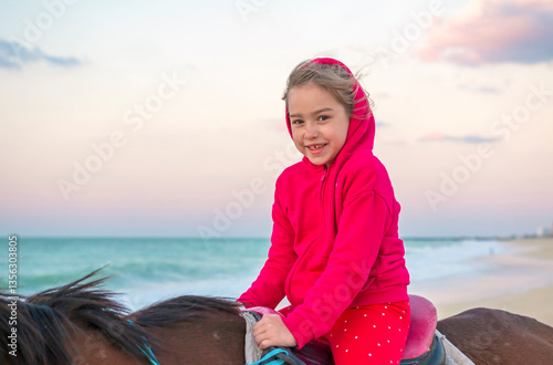Obraz na płótnie A joyful girl smiling while riding a horse on the beach in Sharjah at sunset. The warm golden light of the setting sun reflects on the water, creating a serene and magical moment w salonie