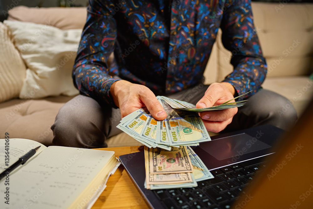 Man holding dollar banknotes, sitting on sofa in front of table with laptop and notebook. Person counting remaining money after paying bills and utility expenses. Personal finance and home budget