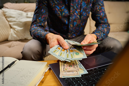 Obraz na płótnie Man holding dollar banknotes, sitting on sofa in front of table with laptop and notebook. Person counting remaining money after paying bills and utility expenses. Personal finance and home budget w salonie