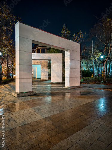 A nighttime scene featuring a modern stone archway in an urban park. the illuminated structure creates a striking contrast against the dark sky, with reflections on the wet pavement adding depth. bare