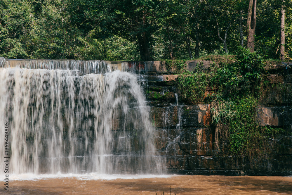 Obraz premium Cascading Waterscape: The raw power and serene beauty of a natural waterfall, cascading gracefully over a tiered stone structure, framed by verdant greenery and lush trees.