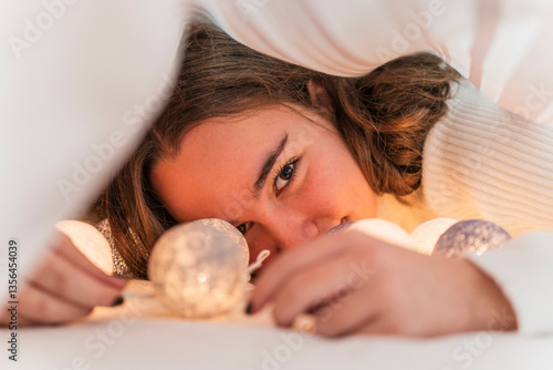 Portrait of teenage girl with chain of lights underneath bedcover
