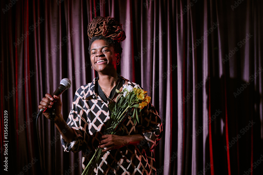 Obraz premium African American performer smiling while holding microphone and bouquet of flowers on stage during performance with long dreadlocks and patterned outfit