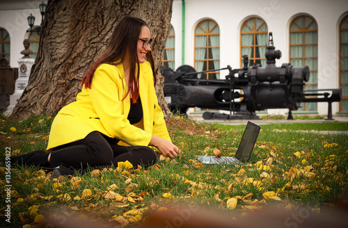 Wallpaper Mural A woman in a yellow jacket works in the park in the fall with a laptop. Woman in classic suit sitting on grass in autumn. Yellow jacket on woman and historical buildings in background. Torontodigital.ca