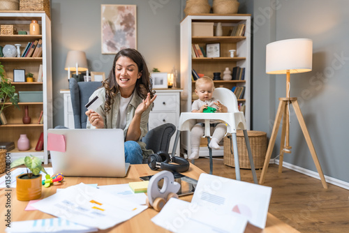 Young woman, mother and business woman, sitting at home online shopping with credit card on laptop computer, while her baby sitting in toddler chair for babies, playing with toy. Mom and kid at home.