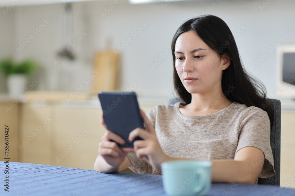 Asian woman reading ebook sitting in a kitchen Asian woman reading ebook sitting in a kitchen