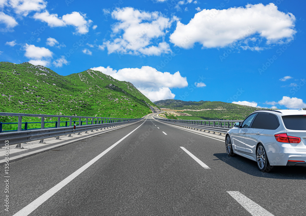A white sedan drives along a modern highway surrounded by lush green mountains, under a bright blue sky with scattered clouds, creating a scenic and peaceful road trip atmosphere.