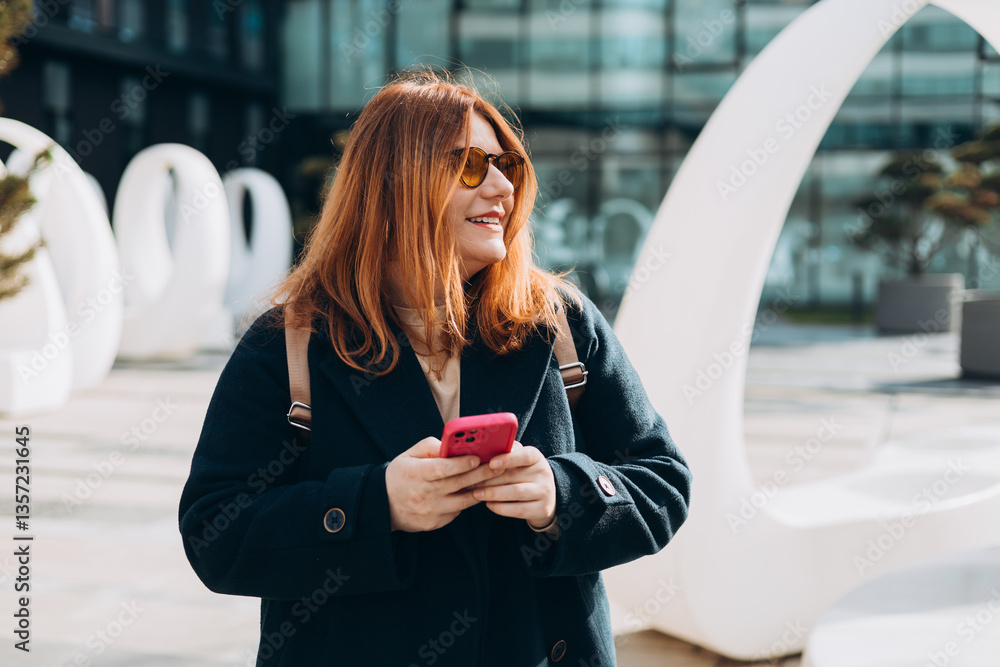 Happy woman chatting on phone and relaxing outdoors. Urban, people concept. Smiling 30s redhead woman text messaging on smart phone at sunny day on the city street, spring time