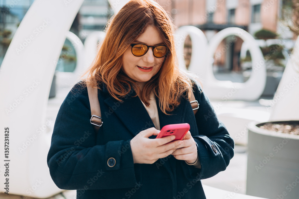 Happy woman chatting on phone and relaxing outdoors. Urban, people concept. Smiling 30s redhead woman text messaging on smart phone at sunny day on the city street, spring time