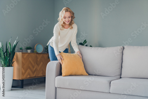 Bilde på lerret Young woman arranging a yellow pillow on a grey sofa in a bright modern living room, showcasing a cozy and relaxed home atmosphere
