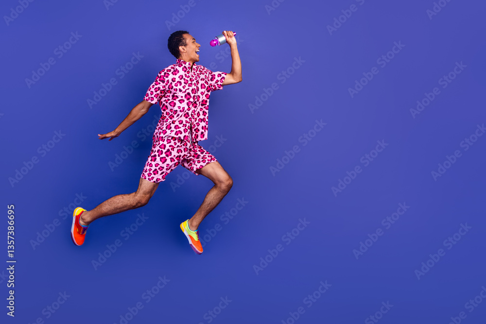 Fototapeta premium Cheerful young man in pink leopard outfit jumping against violet background
