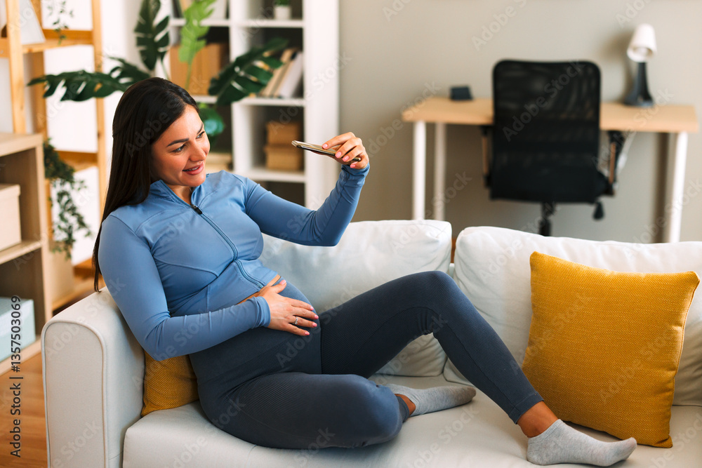 Obraz premium Happy pregnant woman taking a selfie with smartphone while sitting on a comfortable sofa at home