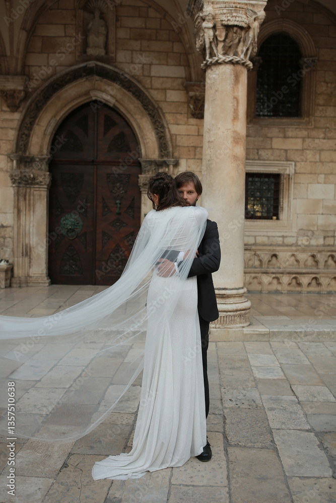 Obraz premium A romantic wedding moment in Croatia. The groom embraces the bride near an ancient building as her veil flows in the wind. The atmosphere is filled with tenderness, love, and historical elegance.