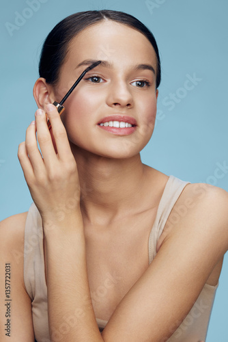 Young woman applying eyebrow gel with a natural smile, showcasing a fresh beauty look against a soft blue background, emphasizing makeup and self care