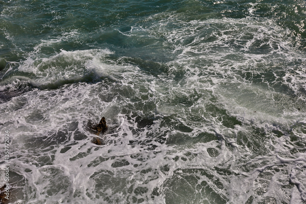 Fototapeta premium Sea foam spreads across uneven surface with visible rocks beneath swirling water and sunlight reflections on green waves; palma de mallorca; turism;