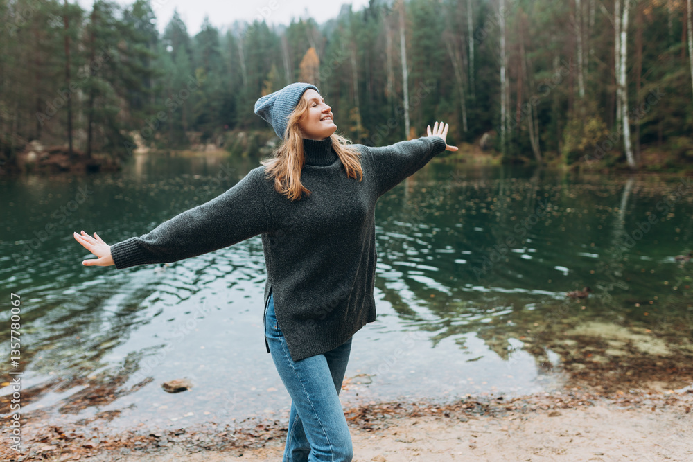 Happy woman raising hands up on the banks of the lake. Young 30s woman hiker in front smiling happy, hiking in woods, warm autumn day.