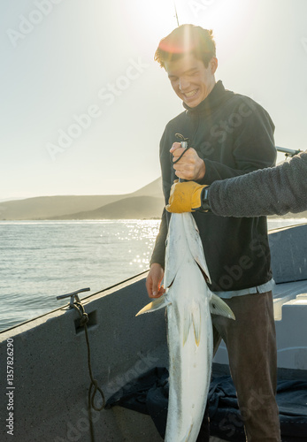 A young fisherman smiles nervously while holding a massive fish he just caught. A helping hand in gloves supports the weight as they pose for the moment.