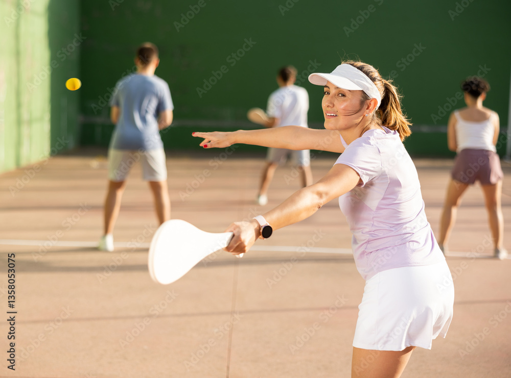 Fototapeta premium Concentrated young hispanic female pelota player swinging wooden paleta to hit ball on outdoor fronton court on sunny summer day. Sport and active lifestyle concept