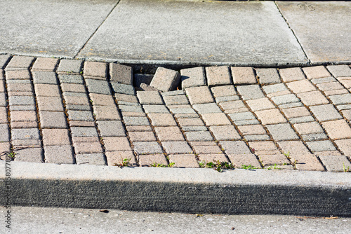 Uneven brick pavement with displaced bricks, adjacent to a concrete sidewalk, surrounded by small patches of grass and weeds