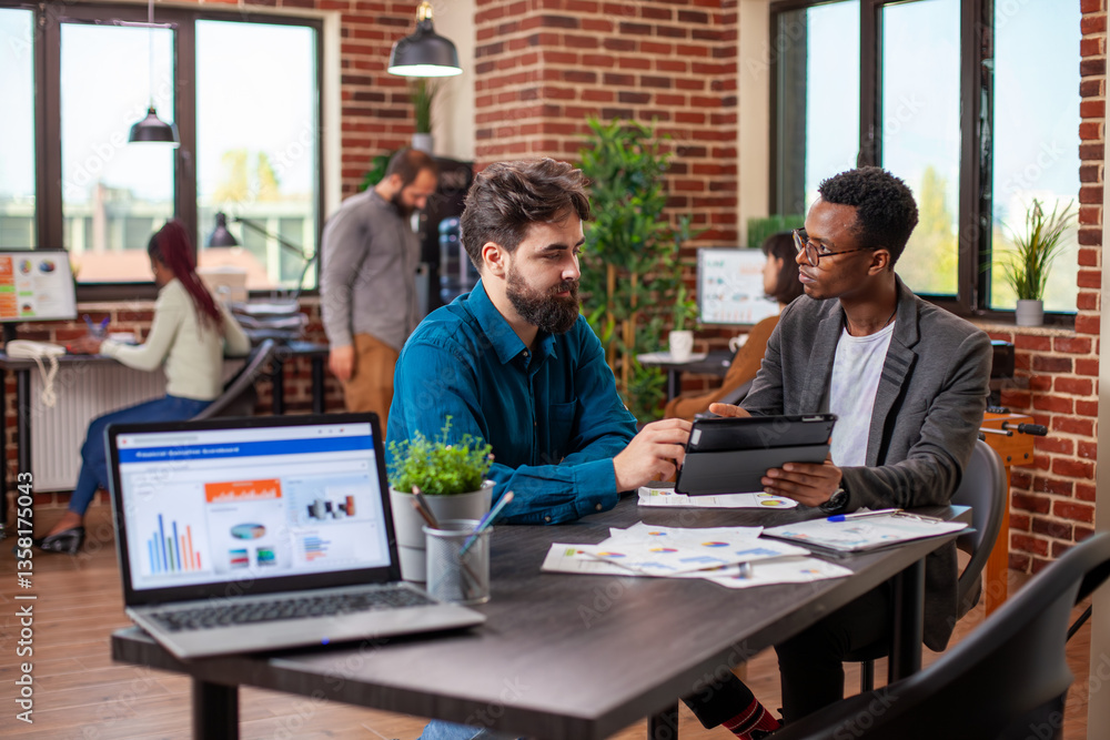 Fototapeta premium Male entrepreneurs brainstorming business ideas and reviewing company data on a tablet in modern workplace. Young men seated at desk with shared device, analyzing annual reports collaboratively.