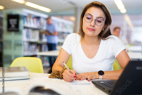 Confident student girl studying in the university library on a laptop, writes an important synopsis in a copybook