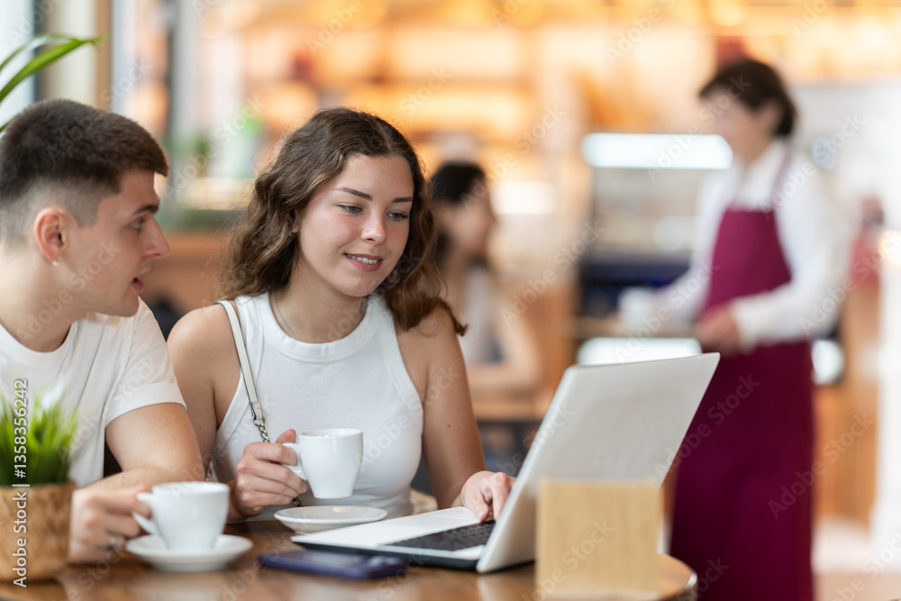 Fototapeta premium Young man and a woman are sitting at a coffee shop table and discussing the details of a project while looking at a laptop. Man with a woman at a business meeting over a cup of hot coffee.