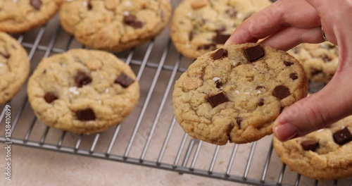 Chocolate chip cookies freshly baked topped with flaky salt on a cooling rack, woman hand taking a cookie