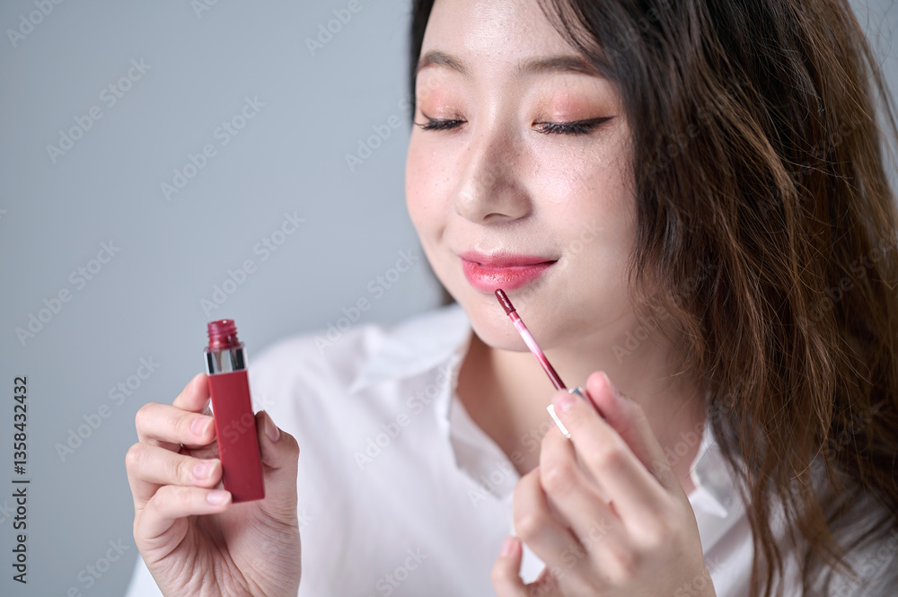 Obraz premium A woman with long hair, wearing a white shirt, is happily applying lip gloss while doing her makeup