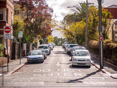 A one-way residential street in an inner suburb of Melbourne, Australia, featuring parked cars along both sides of the road, 'No Entry' signs, and markings indicating traffic direction