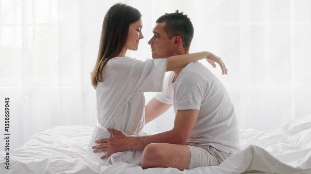A young couple sits on their bed, exchanging loving glances and smiles. The soft morning light filters through white curtains, creating a warm, romantic atmosphere in their home. A young couple sits on their bed, exchanging loving glances and smiles. The soft morning light filters through white curtains, creating a warm, romantic atmosphere in their home.