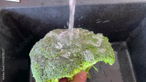 Fresh broccoli being rinsed under running water in a kitchen sink. Water beads up and rolls off the surface, highlighting the vegetable's natural waxy coating that prevents thorough cleaning.