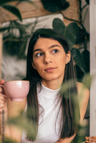 Attractive young dark-haired woman drinking tea from a cup in a room with indoor plants, close-up.