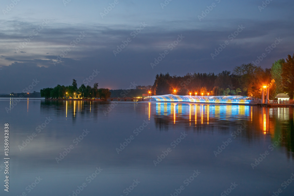 Light fountain with blue and yellow illumination reflecting in the calm water of Ternopil City Pond at twilight, with a small island and trees in the background, Ternopil, Ukraine Light fountain with blue and yellow illumination reflecting in the calm water of Ternopil City Pond at twilight, with a small island and trees in the background, Ternopil, Ukraine