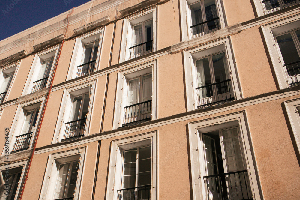 Facade of an apartment block building in old Spanish town. Yellow beige house wall with many windows, balconies with wooden shutters bottom view. Facade of an apartment block building in old Spanish town. Yellow beige house wall with many windows, balconies with wooden shutters bottom view.