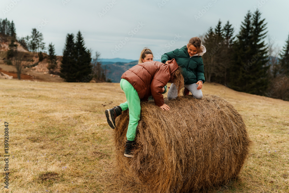 Sisters playing joyfully and climbing on a round hay bale in a picturesque field, relishing their time together on a cloudy day in the serene countryside Sisters playing joyfully and climbing on a round hay bale in a picturesque field, relishing their time together on a cloudy day in the serene countryside