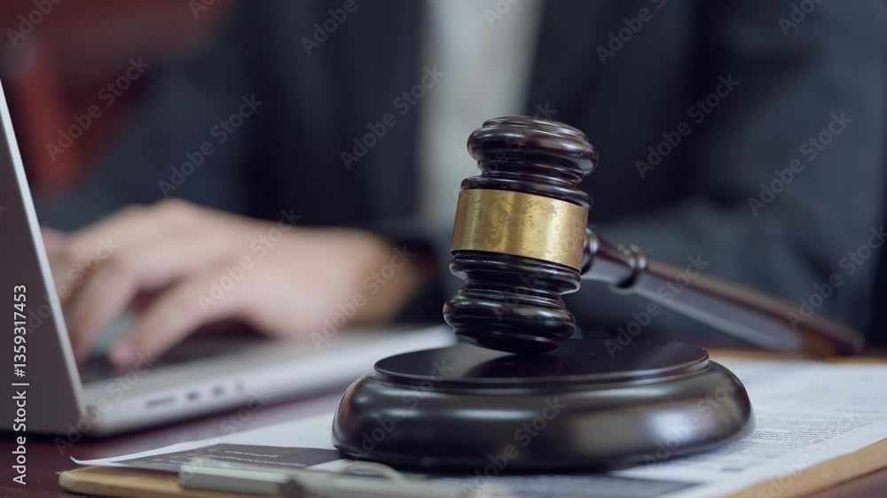 A person is using a laptop computer while a gavel sits on a desk in front of them. The person appears to be working on a legal document or case, as the gavel is a symbol of authority and justice A person is using a laptop computer while a gavel sits on a desk in front of them. The person appears to be working on a legal document or case, as the gavel is a symbol of authority and justice