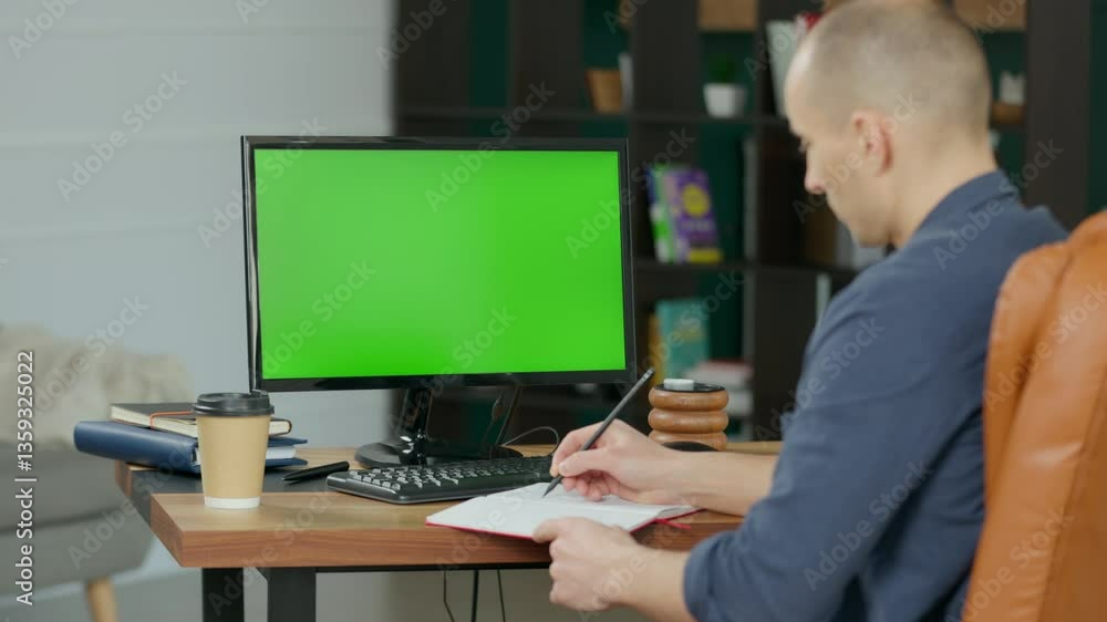 Young Man Writing in Notebook with Pencil near Computer with Green Screen Mock Up Display. Male Manager With Short Hair Wearing Shirt in Modern Home Office Young Man Writing in Notebook with Pencil near Computer with Green Screen Mock Up Display. Male Manager With Short Hair Wearing Shirt in Modern Home Office