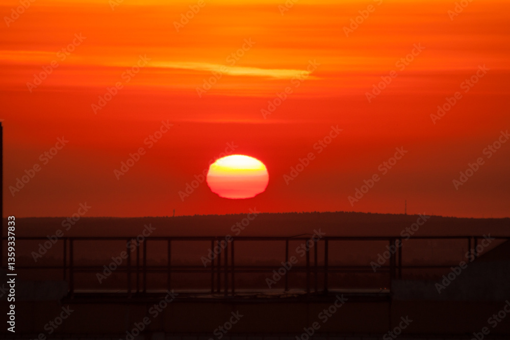 Fototapeta premium View from the roof of a multi-storey brick house in the early morning or late evening to a beautiful bright orange crimson red sunrise or sunset of the heavenly sun from under the hill on which trees.