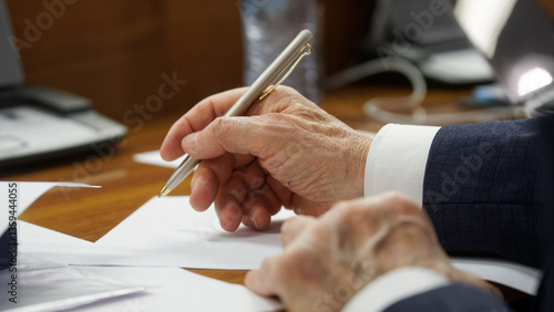 An elderly man's hand carefully signing a formal document with a sophisticated pen. Ideal for illustrating legal, political, or business negotiations and agreements. Photo