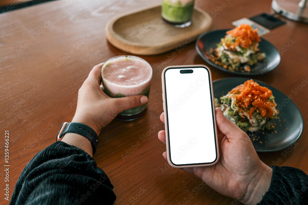 Person holding a smartphone with blank white screen and a colorful layered drink in a cafe, with plates of gourmet food and cozy interior in the background