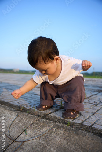 The image shows a young child, possibly a baby or toddler, standing on one leg and playing with a string or cord. The child is wearing a white shirt and appears to be enjoying the activity.