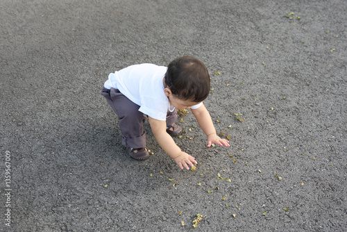 The baby is playing with the leaves on the ground, reaching out to pick them up and play with them. The baby is wearing a white shirt and grey pants while playing outside in the park.