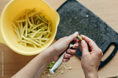 Hands are peeling green beans over a yellow bowl on a wooden table. A board is lying nearby, and the process of preparing vegetables creates an atmosphere of home cooking and care.