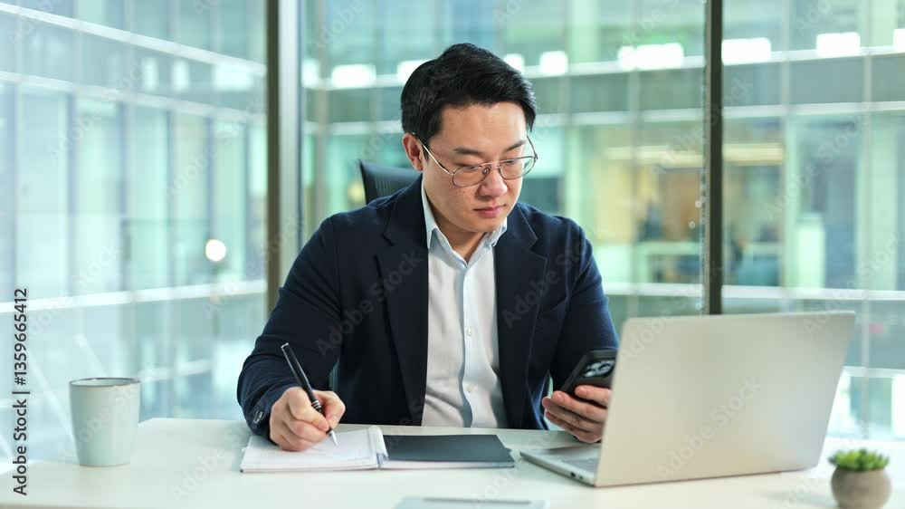 Focused Korean businessman writing notes in notebook while checking smartphone. Adult male entrepreneur working at desk in modern office. Focused Korean businessman writing notes in notebook while checking smartphone. Adult male entrepreneur working at desk in modern office.