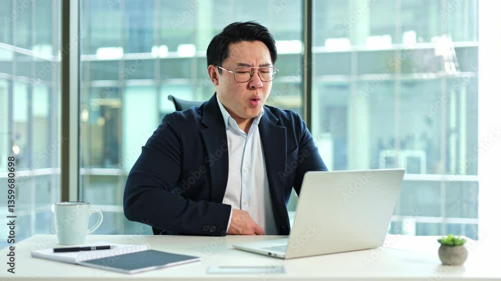 Stressed Asian businessman having a stomach ache in the office. He is sitting at his desk with his hand on his stomach, indicating pain and stress. Stressed Asian businessman having a stomach ache in the office. He is sitting at his desk with his hand on his stomach, indicating pain and stress.