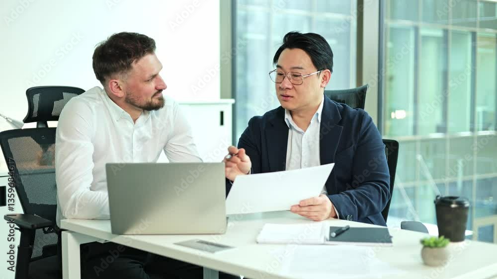 Two businessmen, one Asian and one Caucasian with a beard, review documents together in a modern office using a laptop. Two businessmen, one Asian and one Caucasian with a beard, review documents together in a modern office using a laptop.