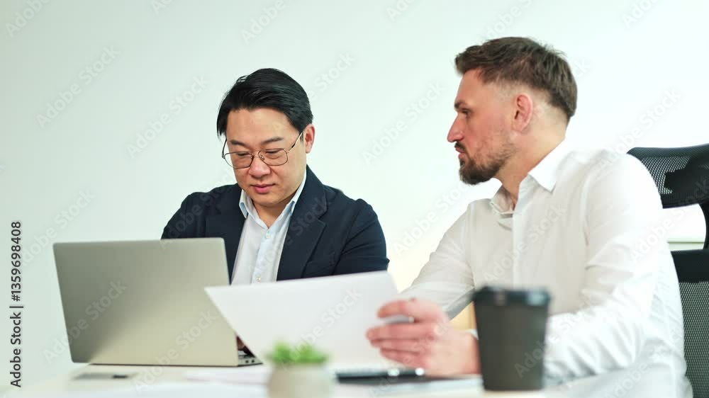 Two diverse businessmen sit in a meeting going over paperwork and discussing what is on a laptop computer. Two diverse businessmen sit in a meeting going over paperwork and discussing what is on a laptop computer.